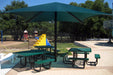 Outdoor picnic area with green tables and benches under a large green Single post Pyramid umbrella, surrounded by trees and playground equipment.