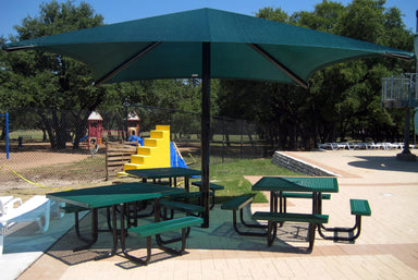 Outdoor picnic area with green tables and benches under a large green Single post Pyramid umbrella, surrounded by trees and playground equipment.