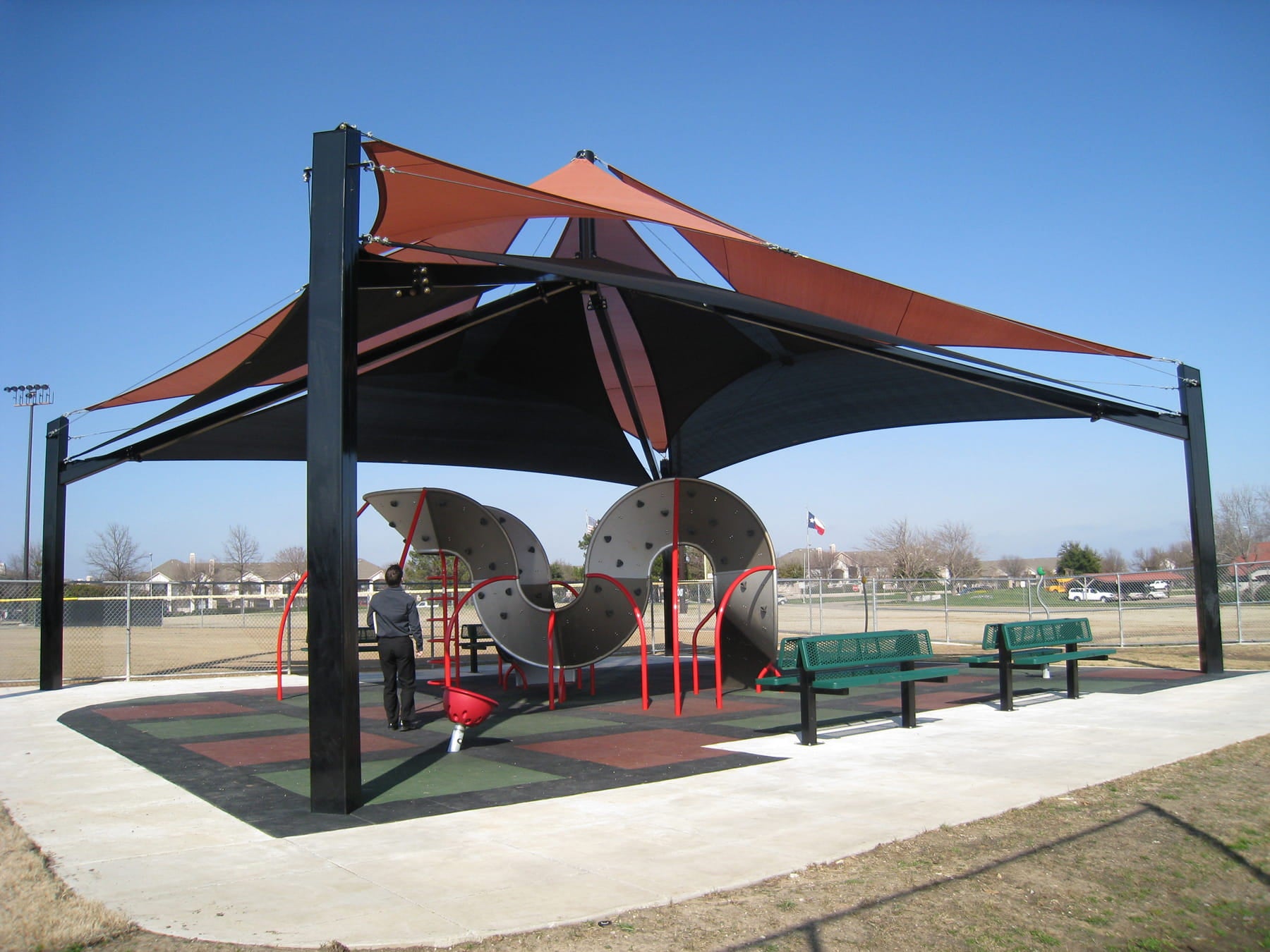 Covered playground area with slides and benches under a large USA Shade Multi-layer Super Span Pyramid Shade structure.