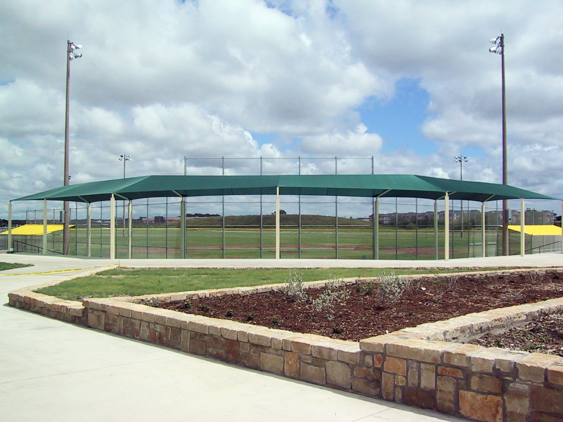 Outdoor sports facility with a green roofed USA Shade Wrap-around Hip and sports field under a cloudy sky.