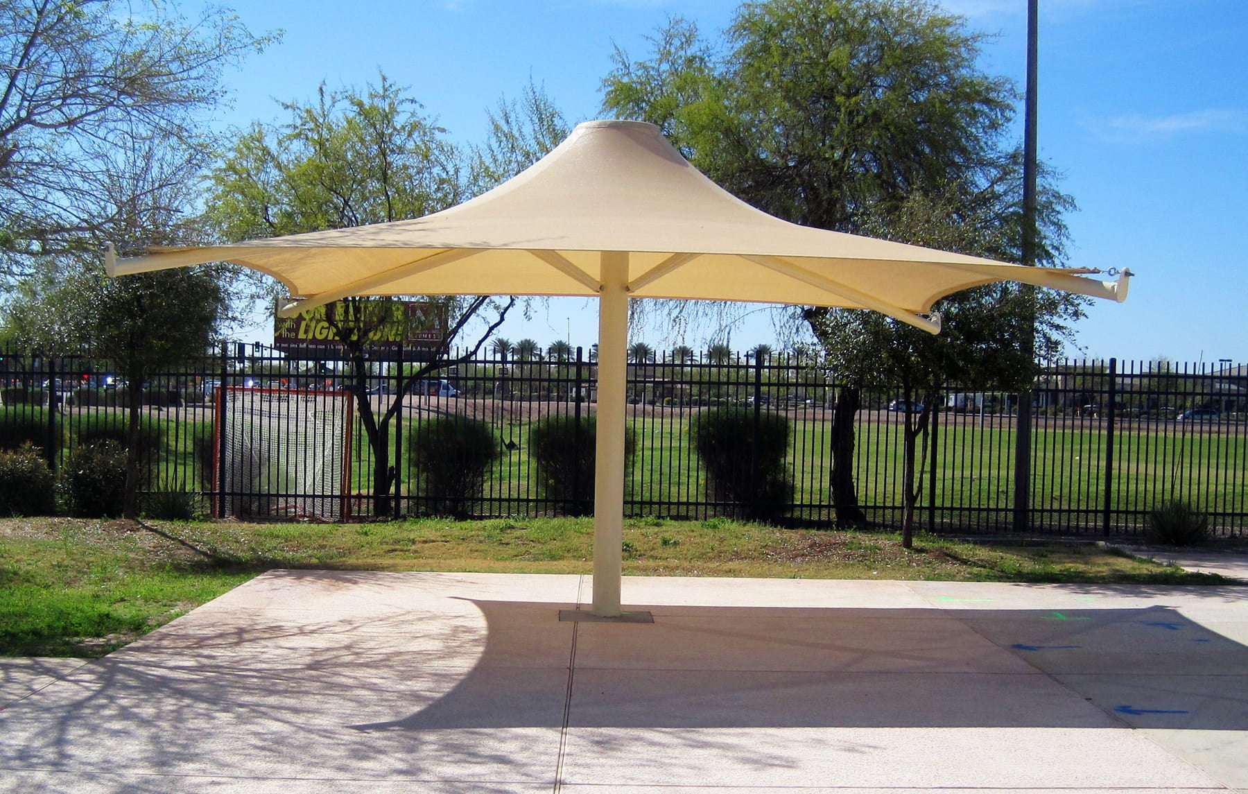 Large beige USA Shade 4-point Twilite Umbrella providing shade in an outdoor setting with trees and a fence in the background.