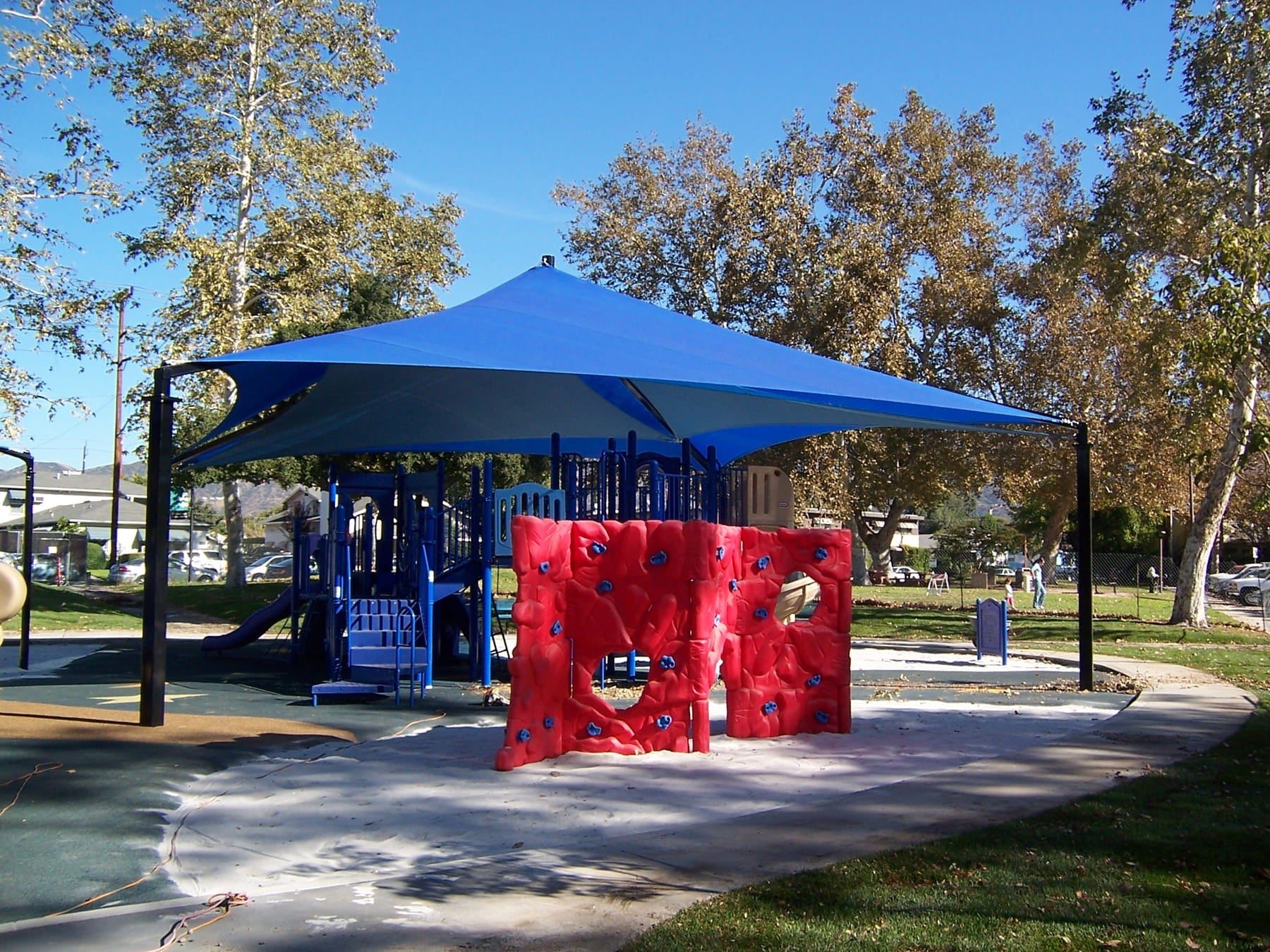 Children's playground with blue USA Shade Mariner Pyramid Shade and red climbing wall in a park setting.