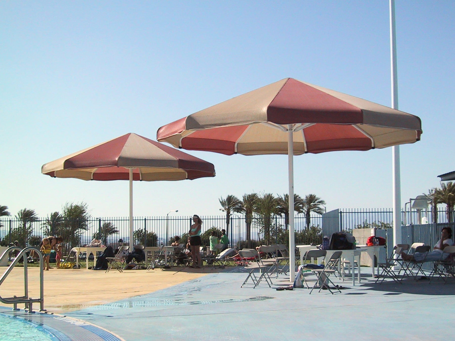 Two large red and beige USA Shade Coolbrellas by a pool with people in the background.