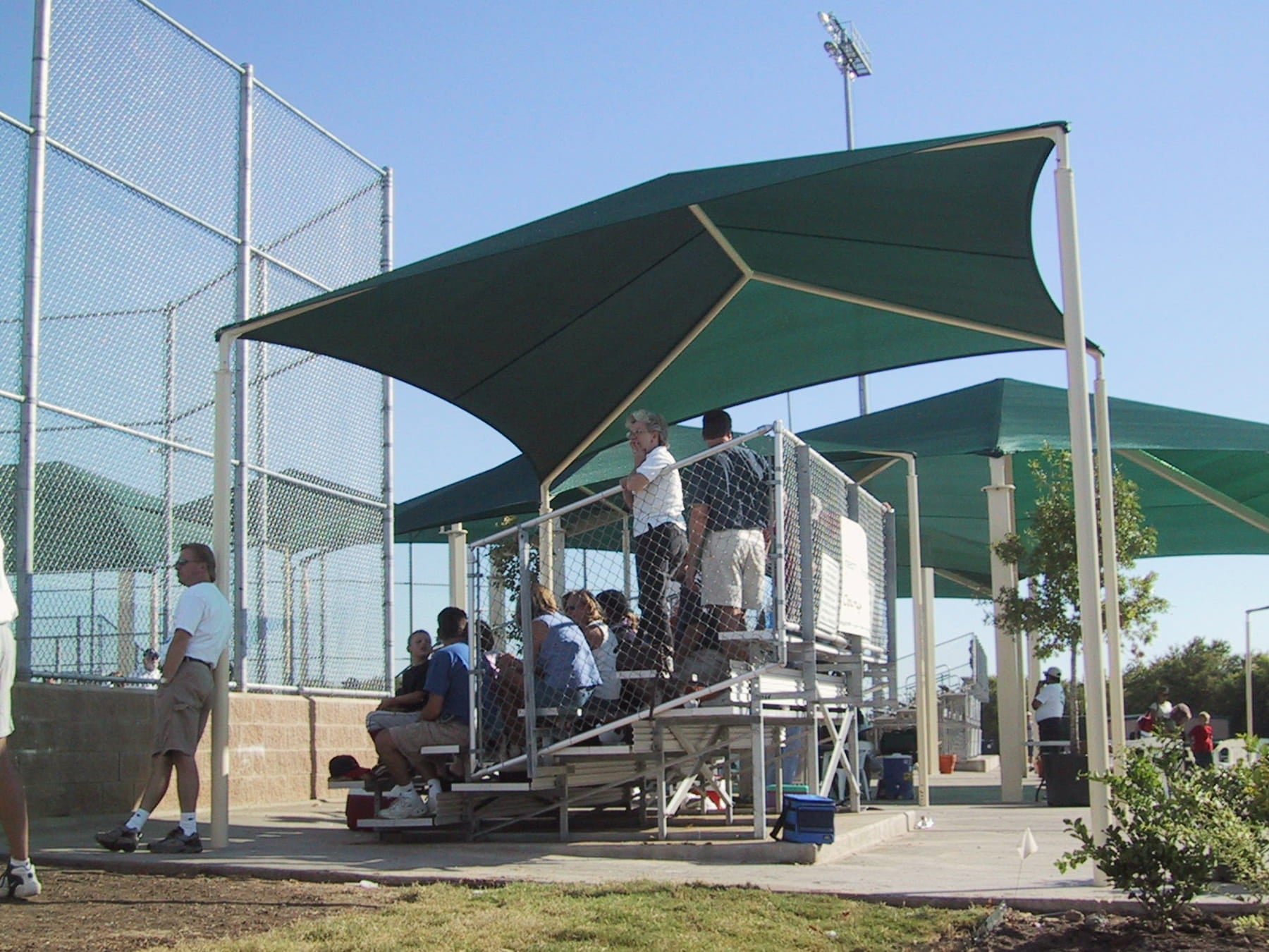 People gathered under a green USA Shade Slant Hip on a sports field with a clear sky.