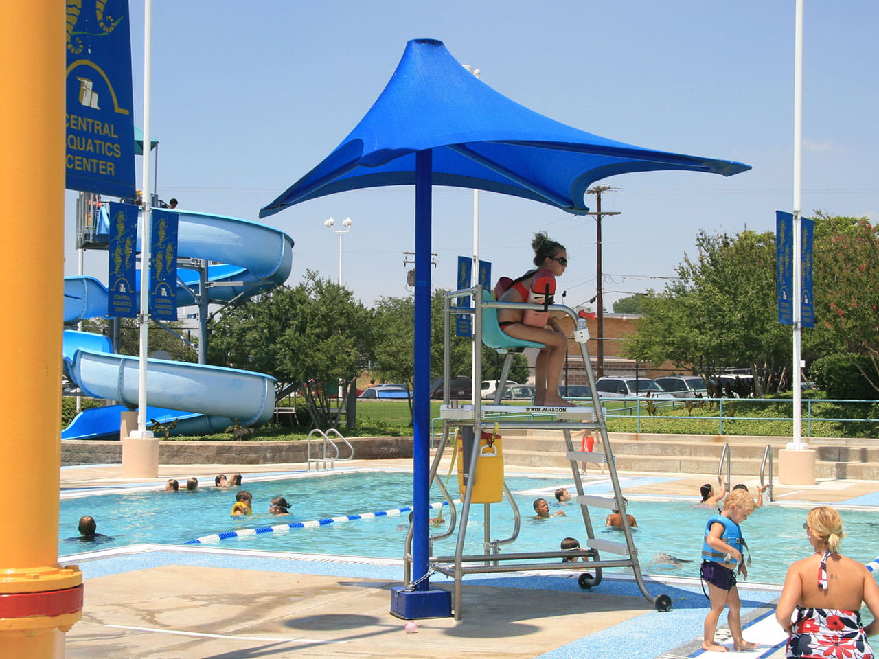 Lifeguard tower with a blue USA Shade Lifeguard Umbrella at a pool with children playing.