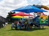People sitting under colorful USA Shade Single Post Pyramid Shades near a playground with slides and inflatable structures.