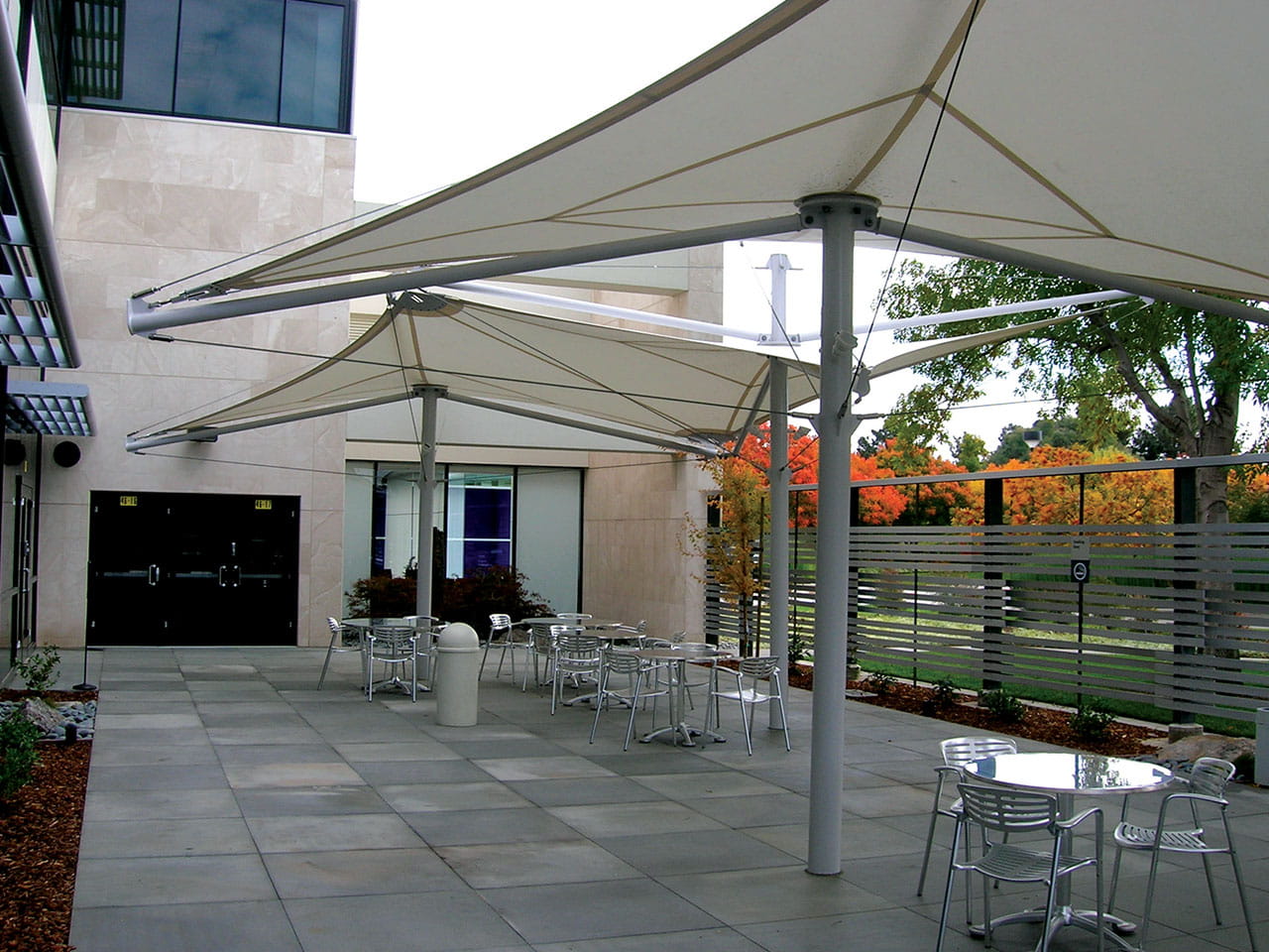 Outdoor patio area with tables and chairs under a large USA Shade Aurora Shade on a sunny day.