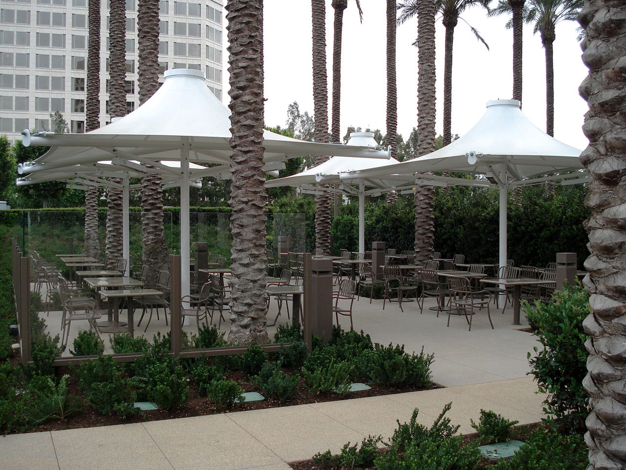 Outdoor seating area with tables, chairs, and USA Shade 4-point Twilite Umbrellas under palm trees and a building.