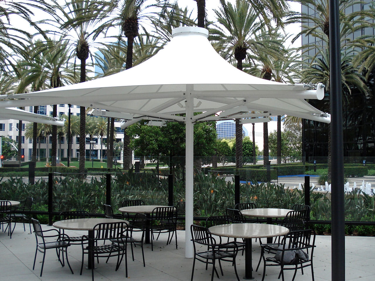 Outdoor seating area with tables and chairs under large USA Shade 4-point Twilite Umbrellas, palm trees, and buildings in the background.