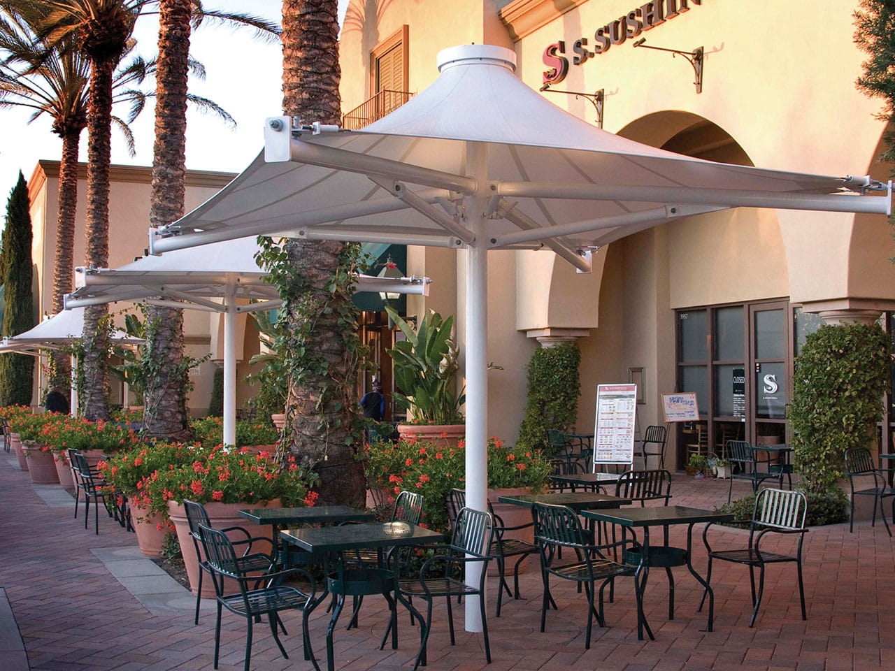Outdoor seating area with tables, chairs, and USA Shade 4-point Twilite Umbrellas in front of a building with 'S Sushi' sign.