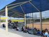 People sitting under a blue USA Shade Wrap-around Hip at a sports facility