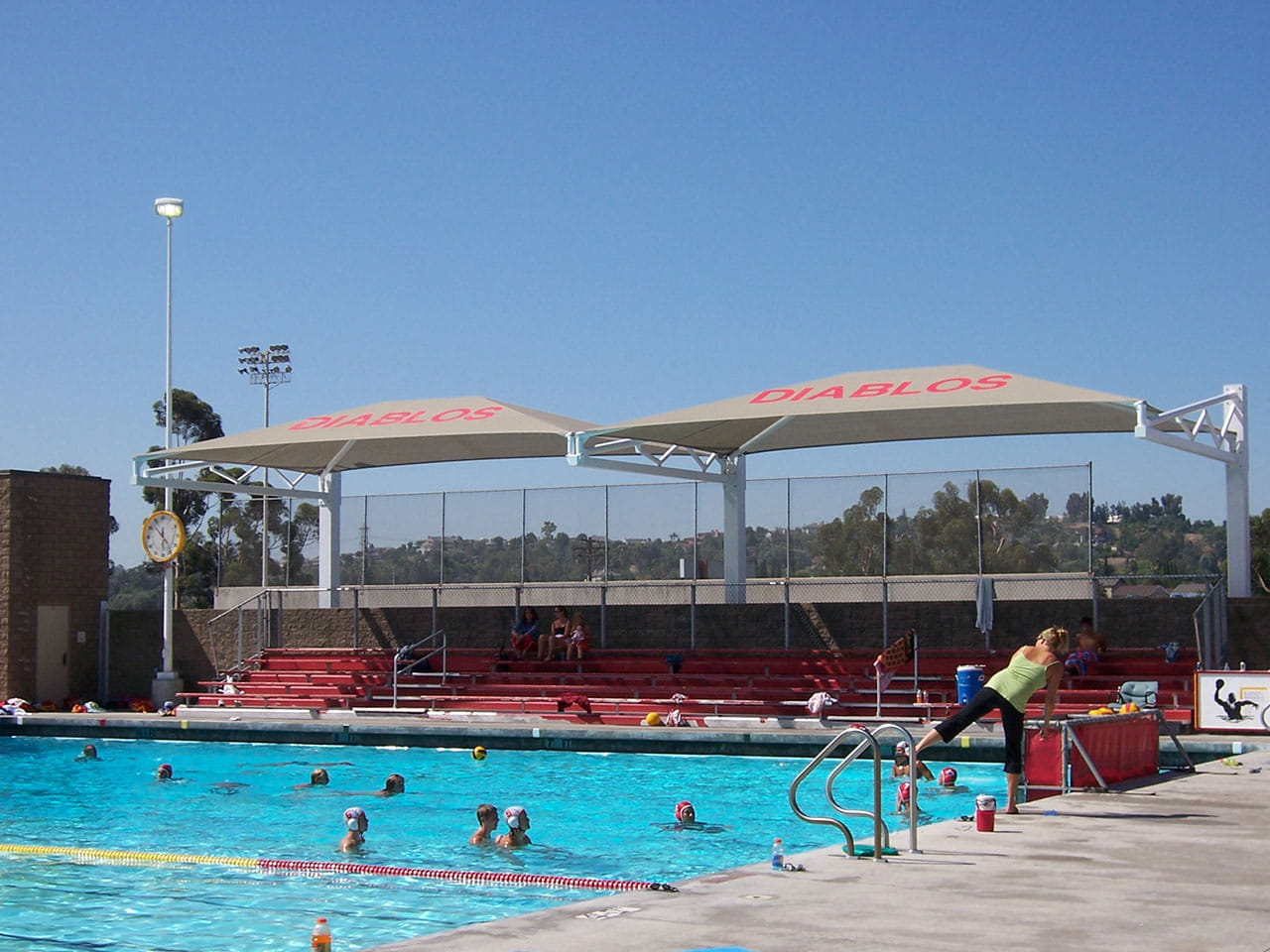 Outdoor pool with people swimming and a large USA Shade Tri-Truss Cantilever with 'Diablos' branding.