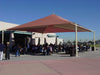 People gathered under a large red and beige USA Shade Mariner Pyramid Shade structure on a clear day.