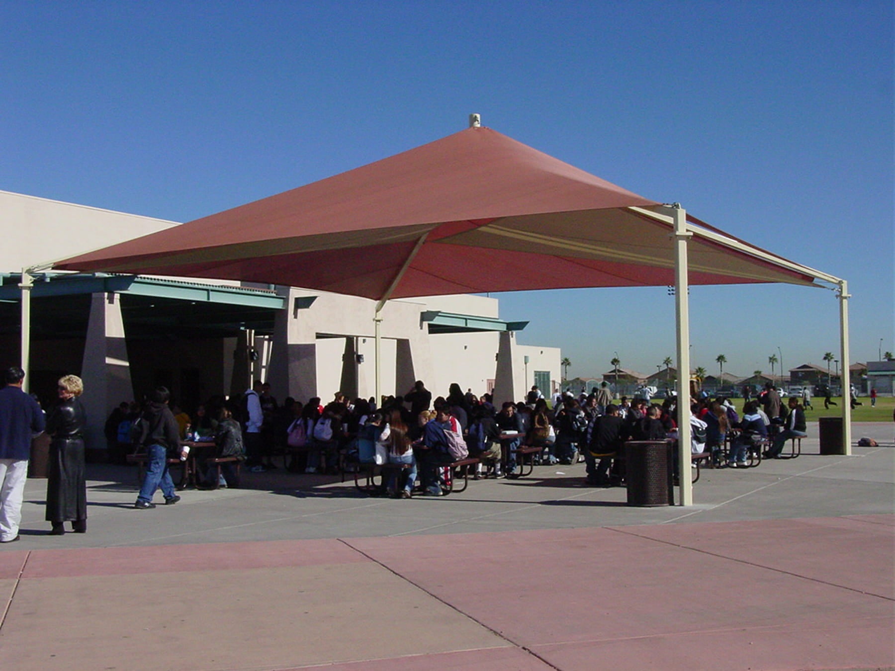 People gathered under a large red and beige USA Shade Mariner Pyramid Shade structure on a clear day.