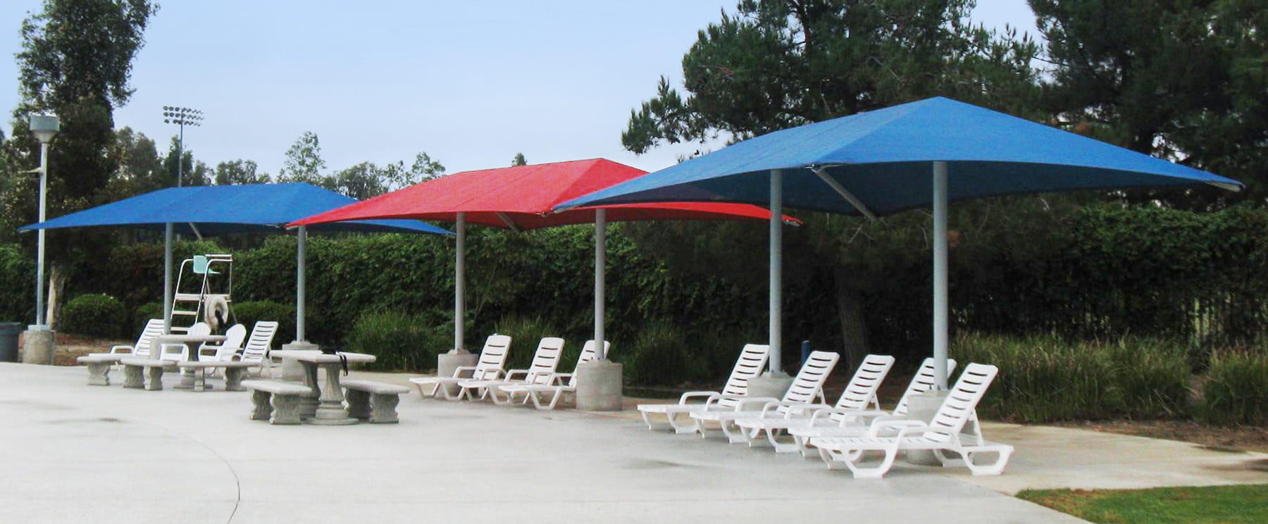 Outdoor seating area with blue and red USA Shade Two Post Hip Shade Structure and white lounge chairs.