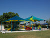 Colorful playground with Teal USA Shade Mariner Pyramid Shade and various play equipment under a clear blue sky.