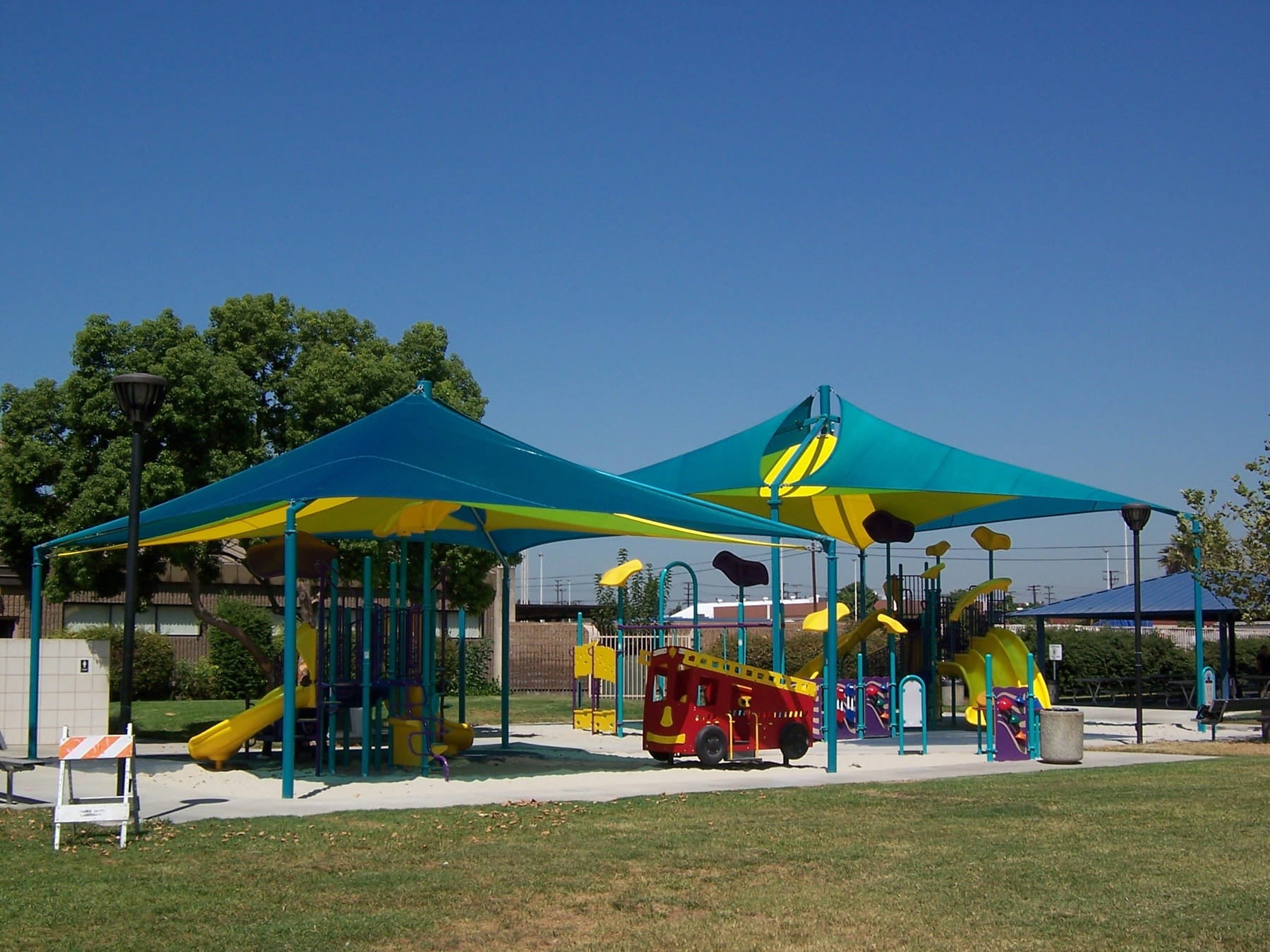 Colorful playground with Teal USA Shade Mariner Pyramid Shade and various play equipment under a clear blue sky.