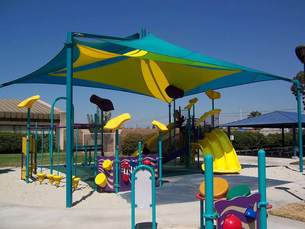 Children's playground with colorful equipment under a large USA Shade Mariner Pyramid Shade structure on a clear day.