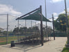 Empty bleachers under a green Cantilever Hypar Sail Shade on a sports Baseball field with trees and sky in the background.