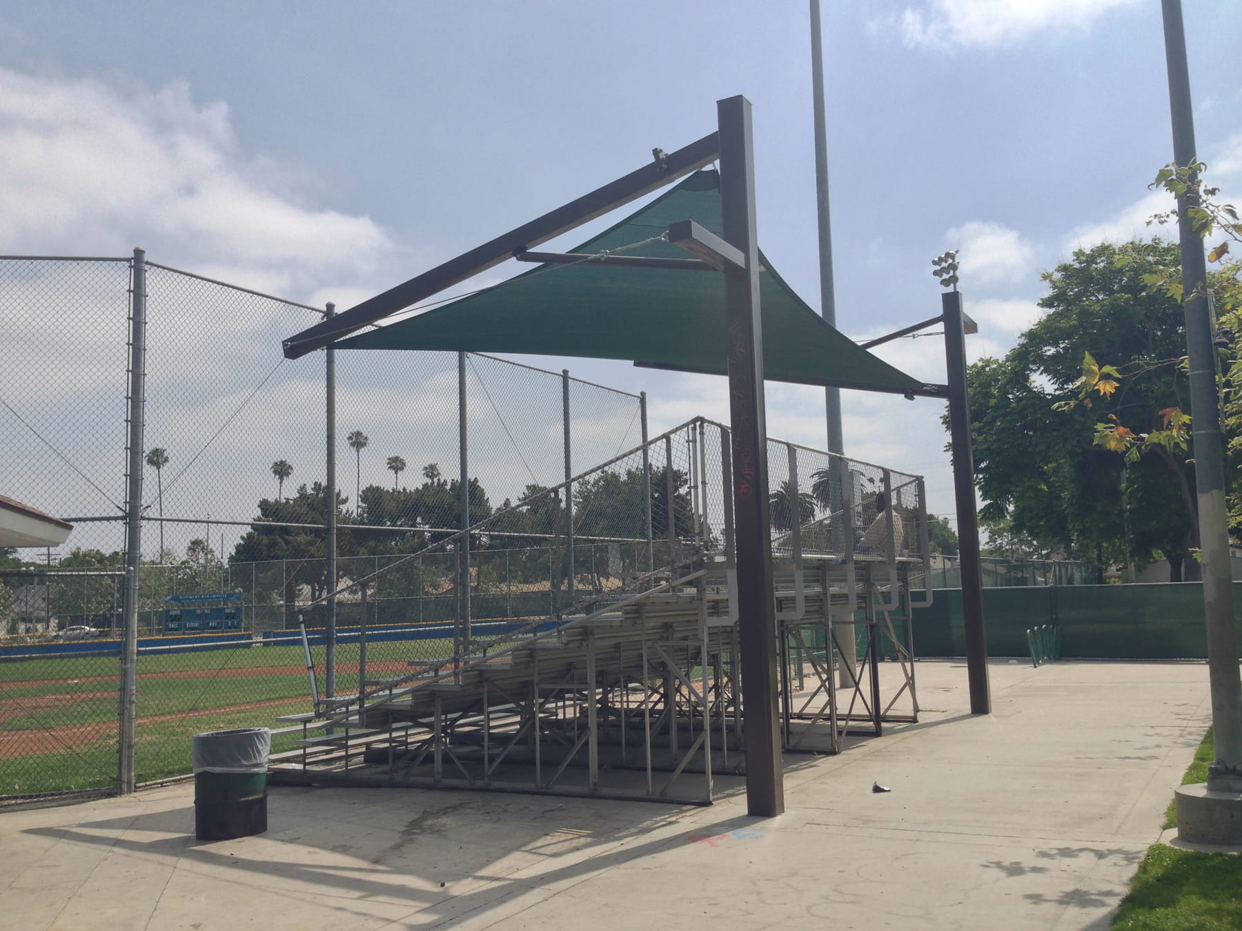 Empty bleachers under a green Cantilever Hypar Sail Shade on a sports Baseball field with trees and sky in the background.