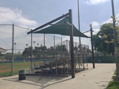 Empty bleachers under a green Cantilever Hypar Sail Shade on a sports Baseball field with trees and sky in the background.