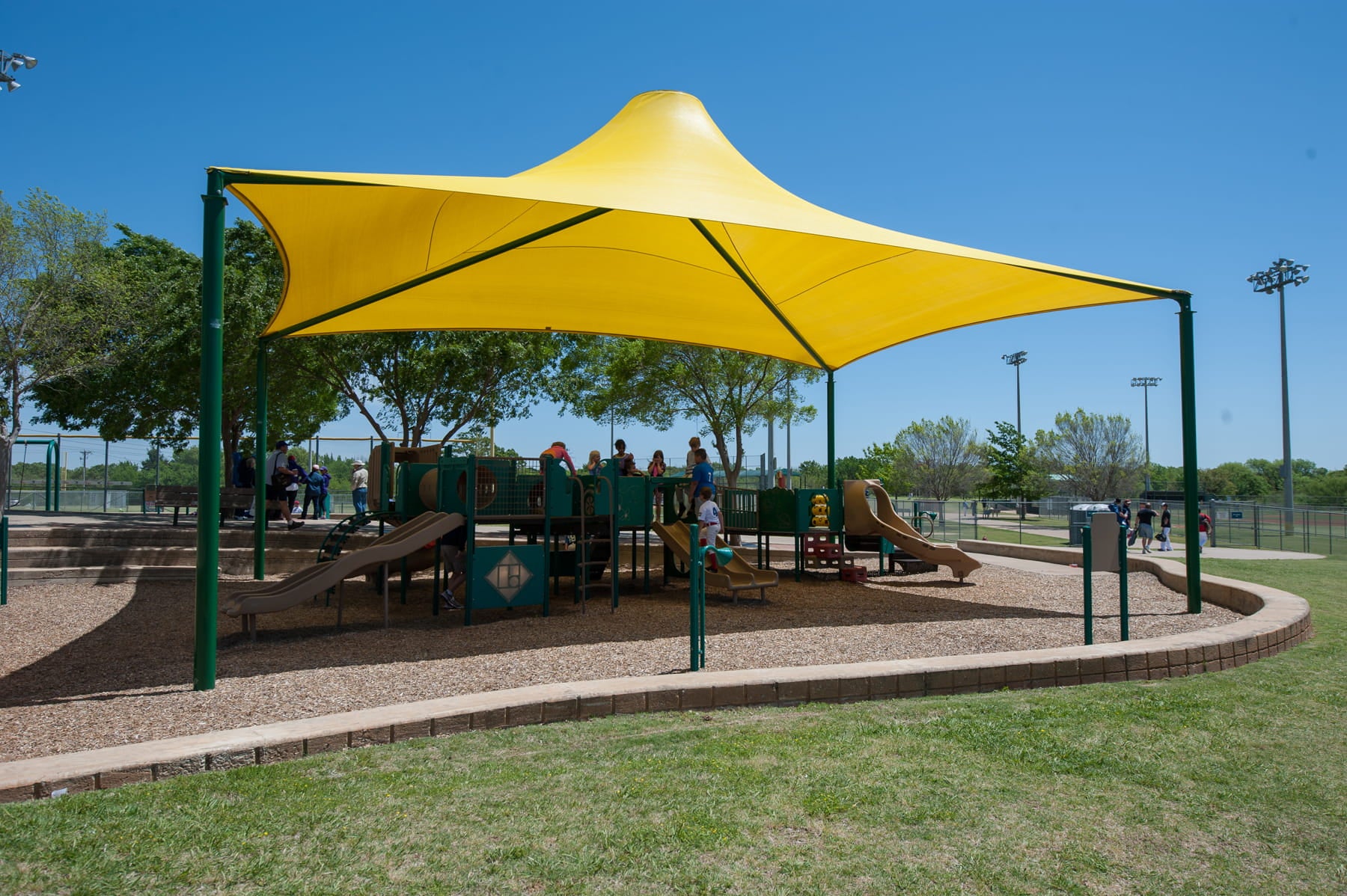 Yellow USA Shade Sahara Shade structure at a playground with children playing underneath on a sunny day.