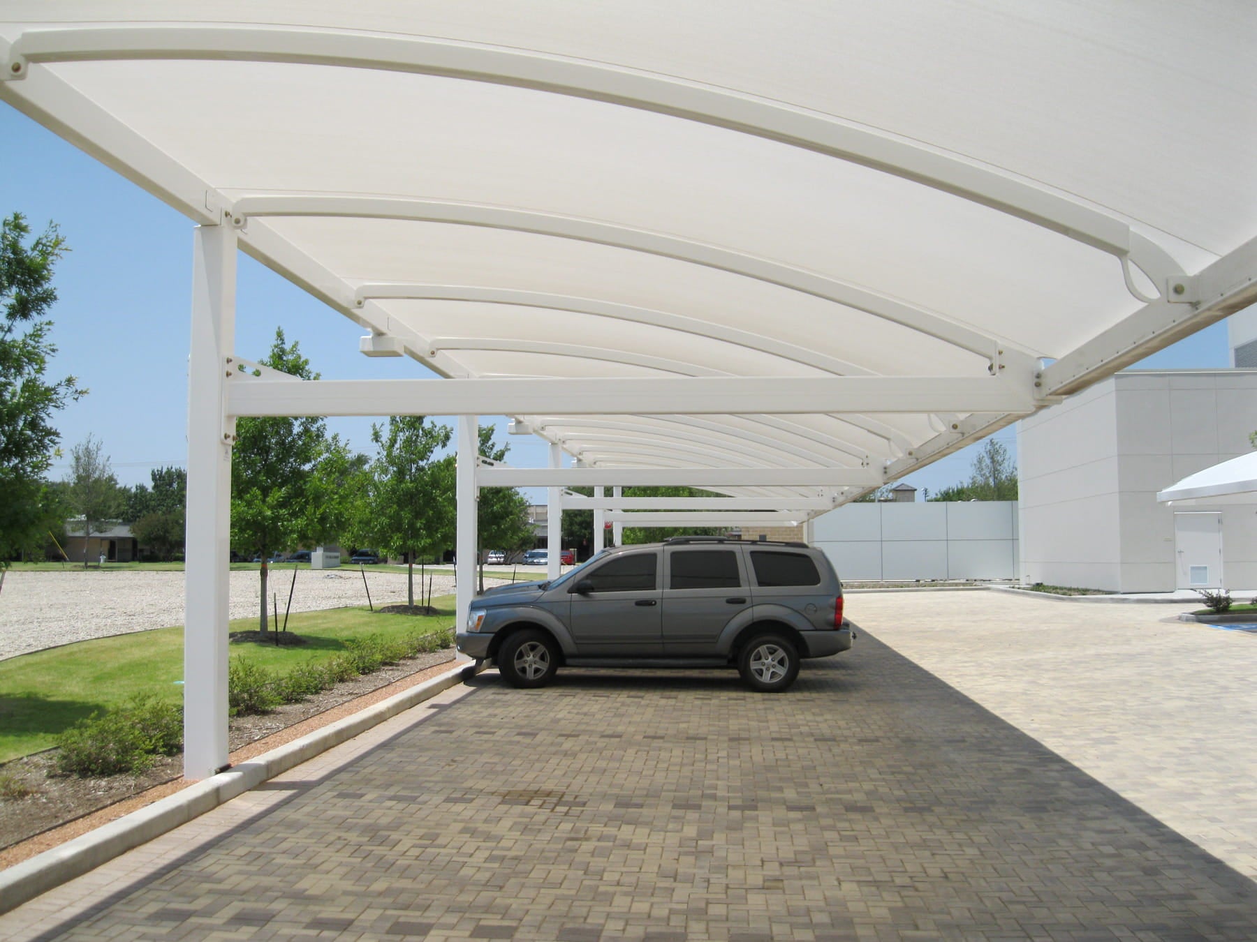 Car parked under a white USA Shade Full Arch Cantilever with trees and a building in the background