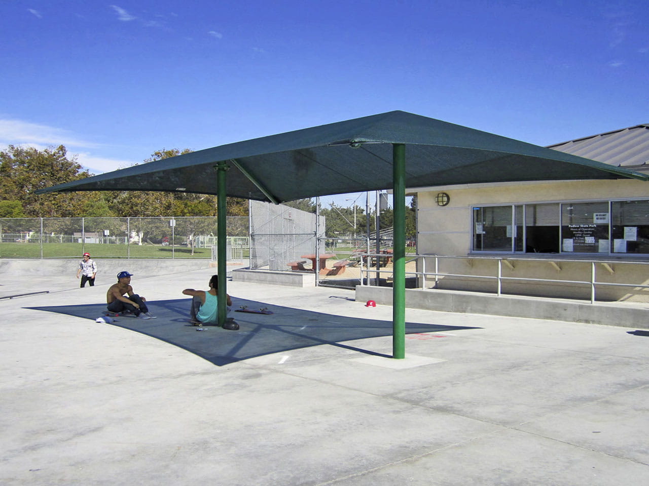 People sitting under a green USA Shade Two Post Hip Shade Structure in an outdoor area with a building in the background.