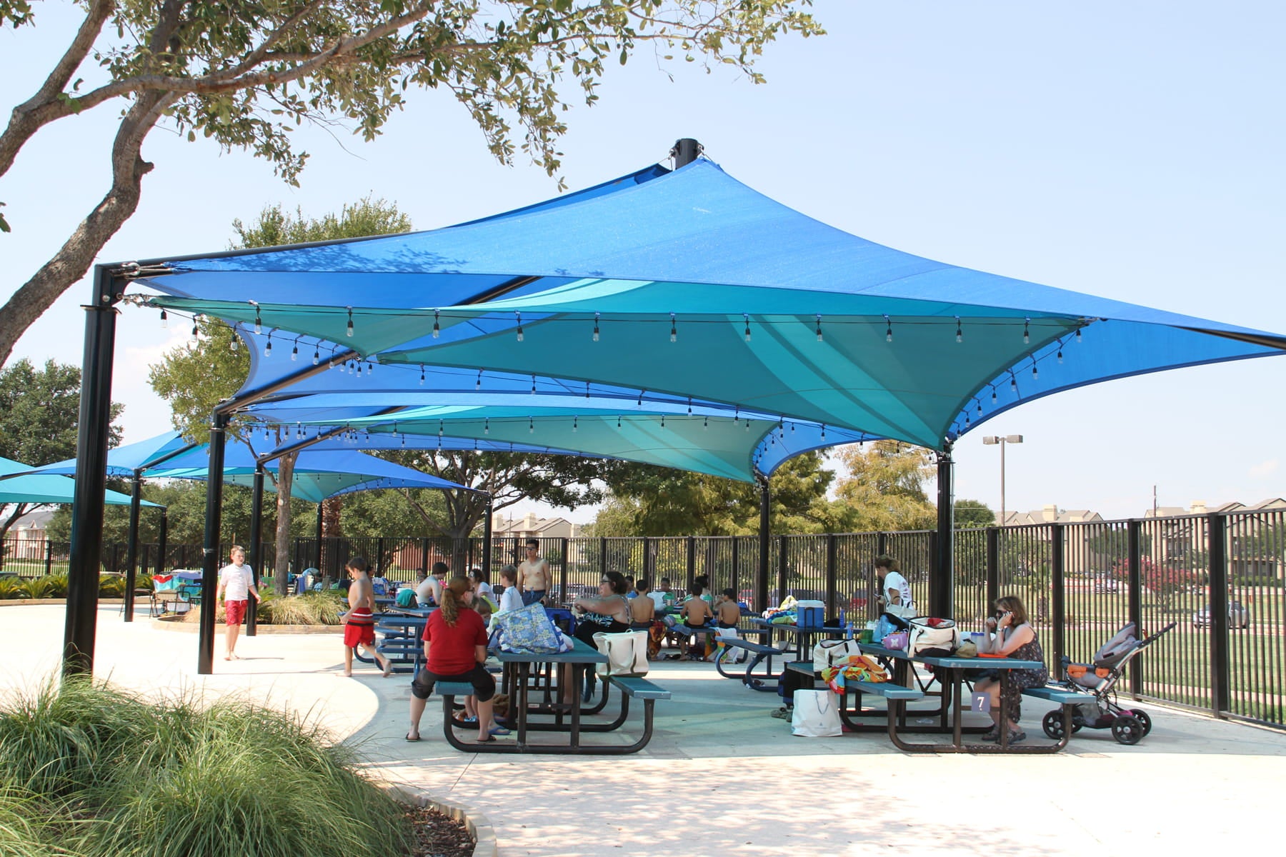 Outdoor shaded area with people sitting at picnic tables under large blue USA Shade Mariner Pyramid Shade.
