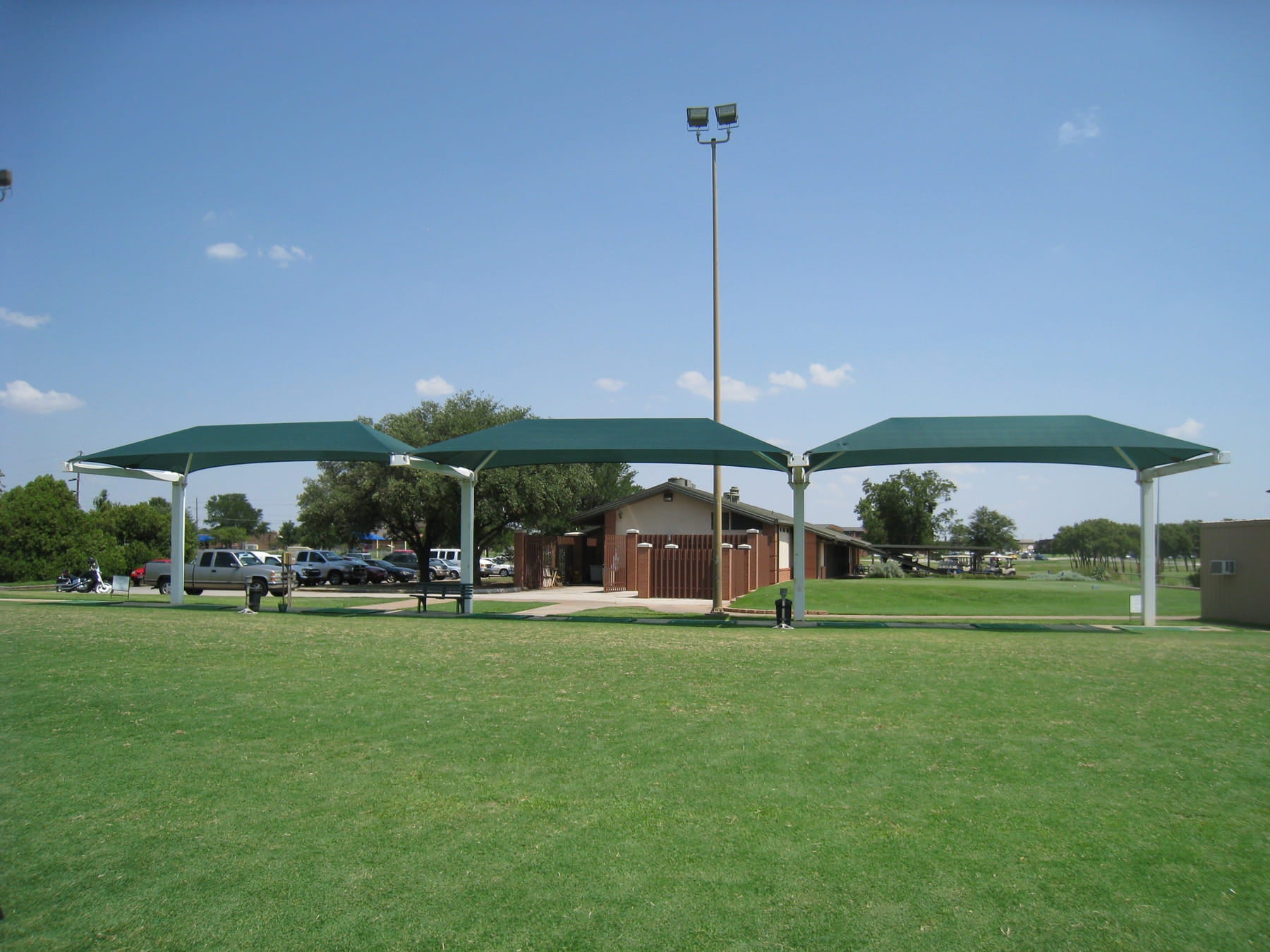 Three green USA Shade Full Hip Cantilever Shades on a grassy field with vehicles and a building in the background.