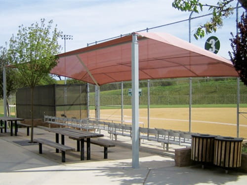 Outdoor seating area with red USA Shade Slant Hip structure, tables, and benches near a sports field.