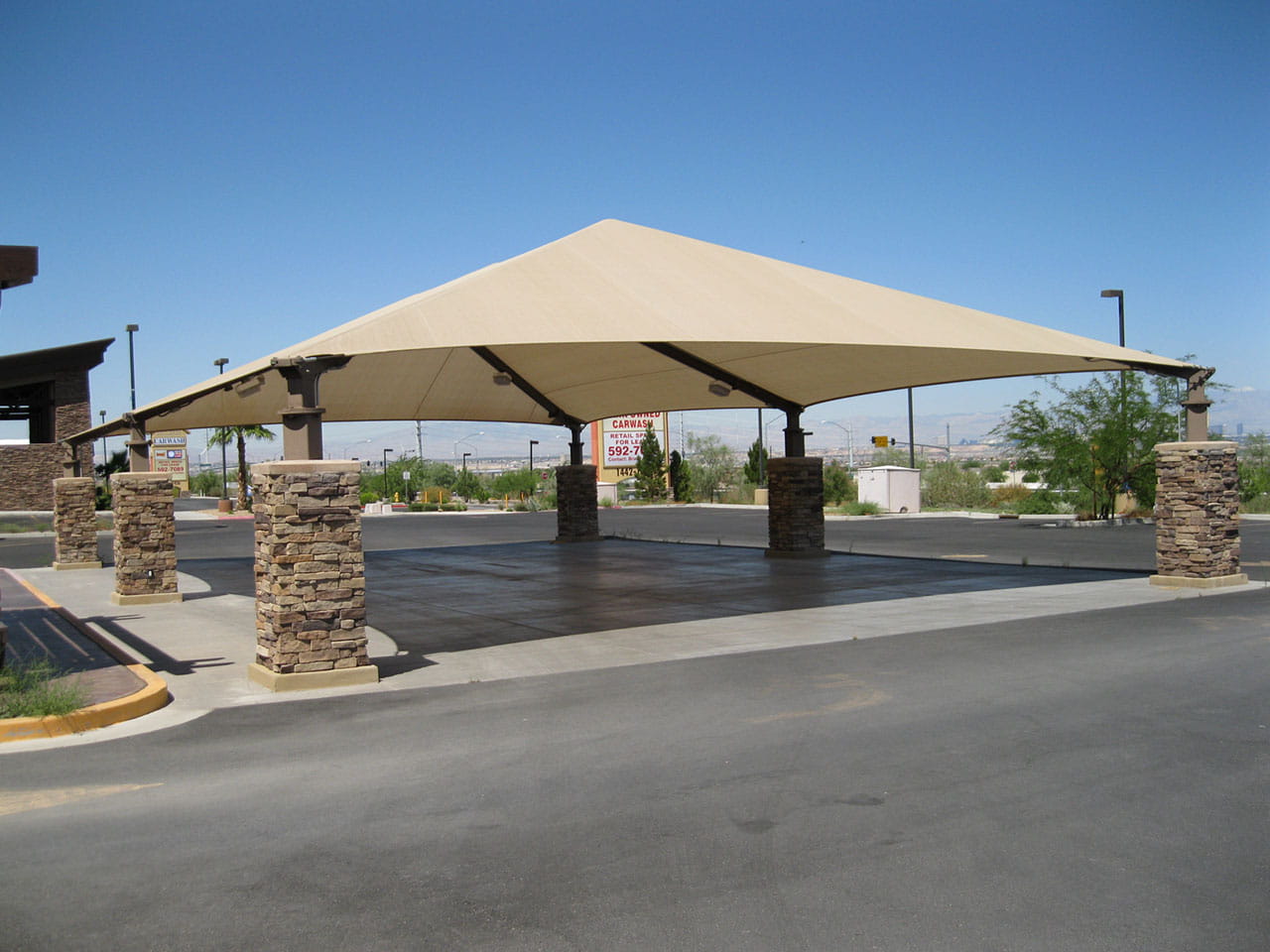 Large beige USA Shade Super Span 6-Post Hip shade with stone columns in a parking lot.