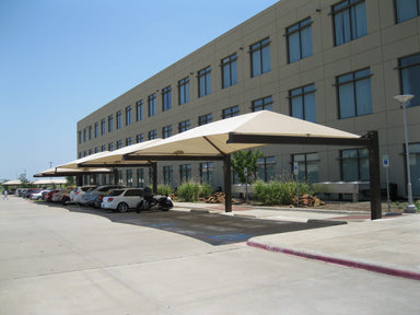 Carport with beige USA Shade Full Hip Cantilever Shades in front of a building with large windows on a clear day.