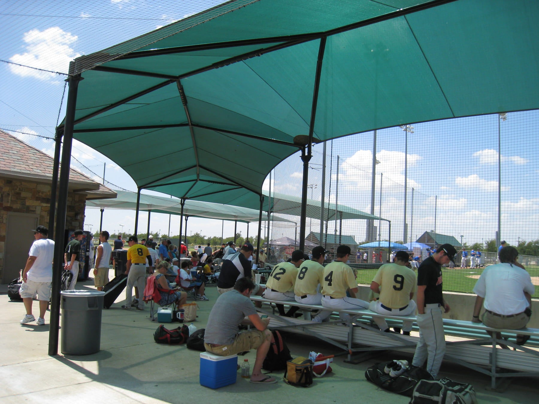 Baseball players and staff in a dugout under a green USA Shade Wrap-around Hip on a sunny day.