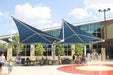 Outdoor area with blue USA Shade Wave Shades, people sitting at tables, and a fountain in front of a building.