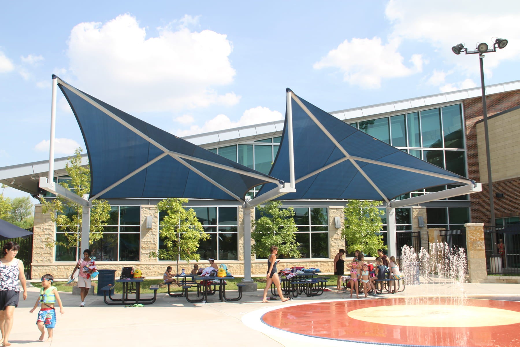 Outdoor area with blue USA Shade Wave Shades, people sitting at tables, and a fountain in front of a building.