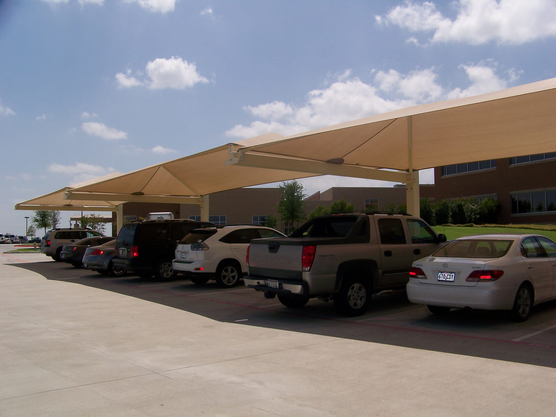 Row of cars parked under large beige USA Shade Full Hip Cantilever Shades on a sunny day.