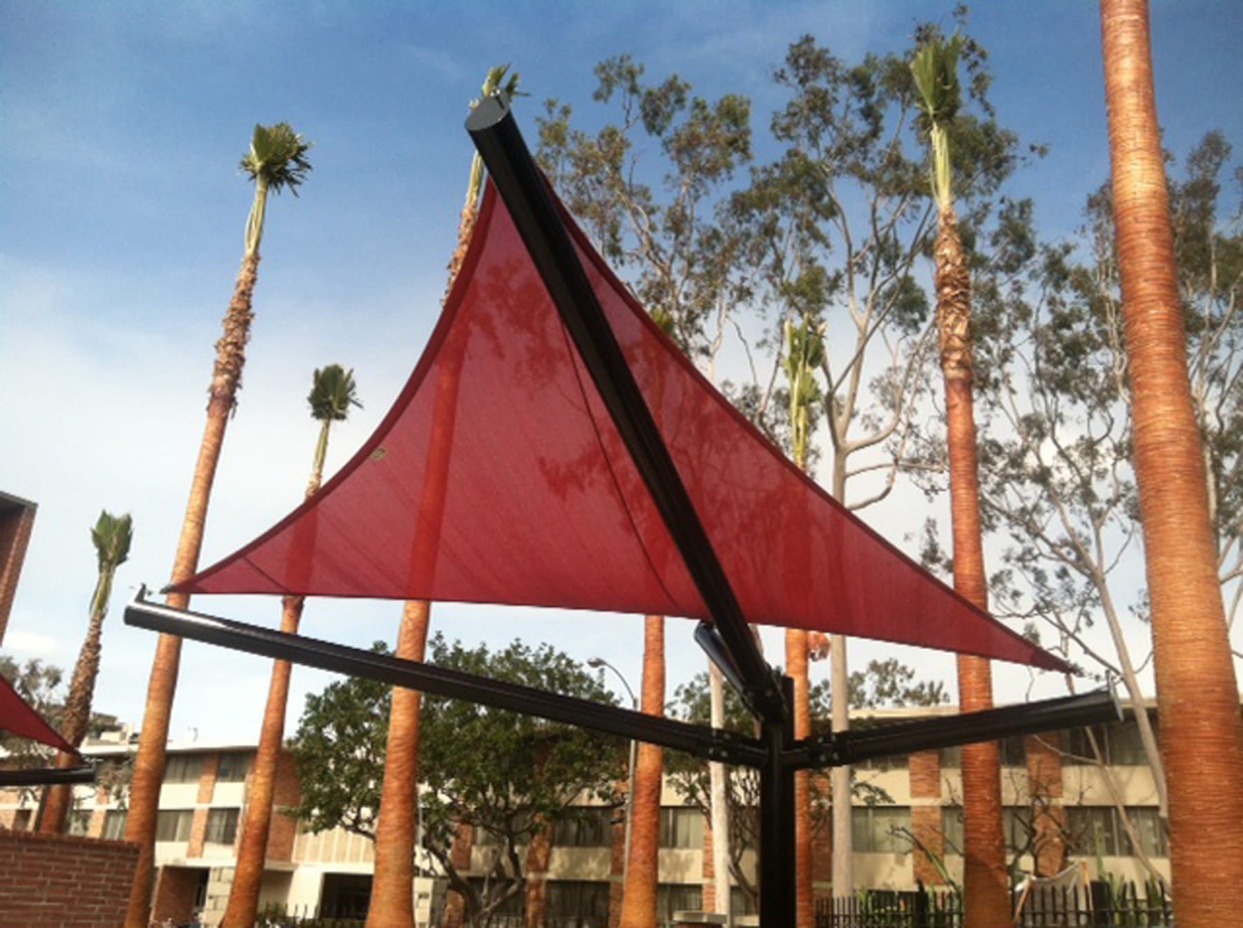 Red USA Shade Arbor Shade sail with black supports against a backdrop of palm trees and buildings.