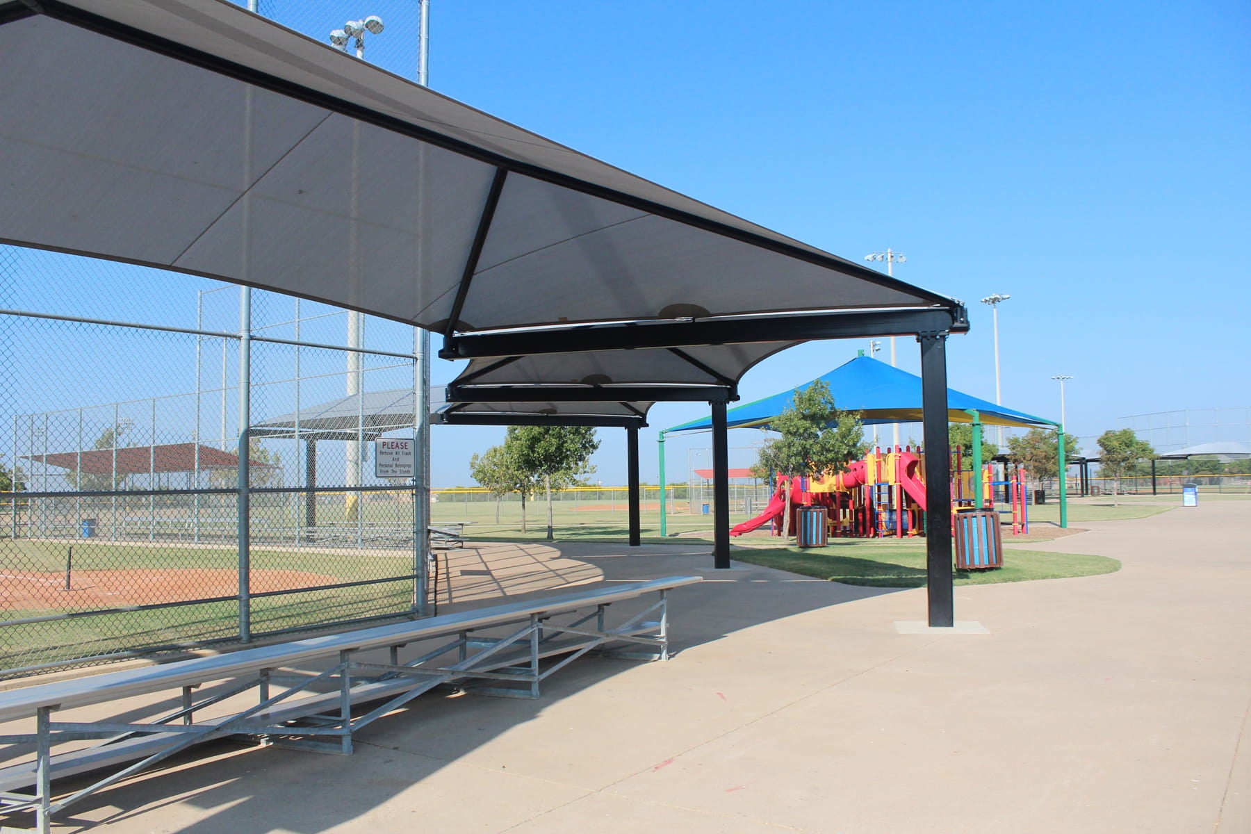 USA Shade Wrap-around Hip covering seating area with bleachers at a sports field, featuring a playground in the background.