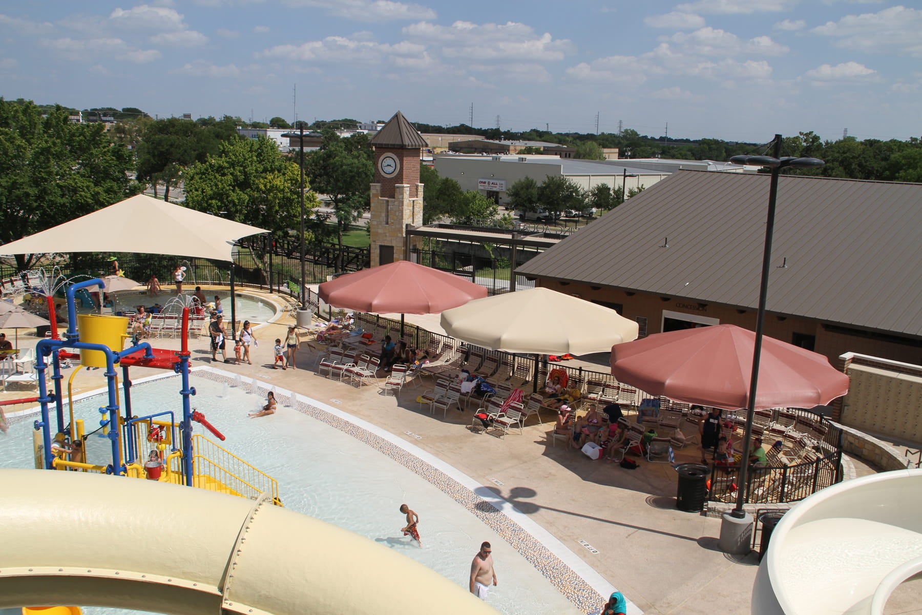 Outdoor pool area with slides, USA Shade Coolbrellas, and people near a clock tower.